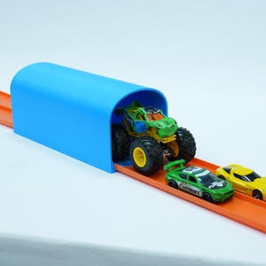 Pu&ograve; includere: Un tunnel di plastica blu con un monster truck e due macchinine su una pista arancione. Il monster truck &egrave; verde e giallo, e le macchinine sono verdi e gialle. La pista &egrave; su sfondo bianco.