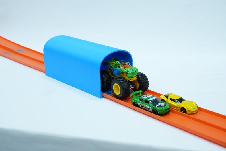 Pu&ograve; includere: Un tunnel di plastica blu con un monster truck e due macchinine su una pista arancione. Il monster truck &egrave; verde e giallo, e le macchinine sono verdi e gialle. La pista &egrave; su sfondo bianco.