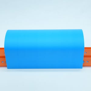 Pu&ograve; includere: Un pezzo di plastica curvo blu brillante &egrave; posizionato sopra due piste per auto giocattolo arancioni. Le piste sono parallele e hanno una fessura centrale. Lo sfondo &egrave; bianco.