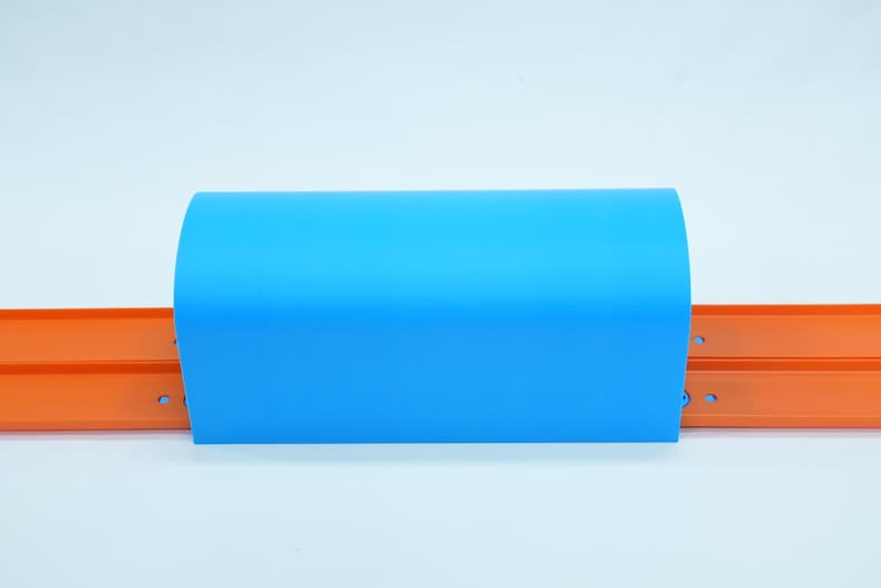 Pu&ograve; includere: Un pezzo di plastica curvo blu brillante &egrave; posizionato sopra due piste per auto giocattolo arancioni. Le piste sono parallele e hanno una fessura centrale. Lo sfondo &egrave; bianco.