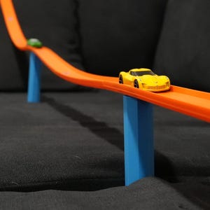 May include: A yellow toy car on an orange track, supported by blue pillars. The track curves upwards, suggesting a race track or play set. A green ball is also on the track. The background is a dark gray surface.