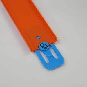 May include: An orange plastic toy track segment with a blue connector. The track has a raised edge and a small blue button. The connector has a circular design with the letters "ABC".
