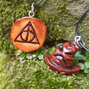 May include: Two handmade ceramic necklaces on a mossy green background. One necklace features a brown pendant with a dark brown triangle and circle design. The other necklace features a brown pendant shaped like a wizard's hat.