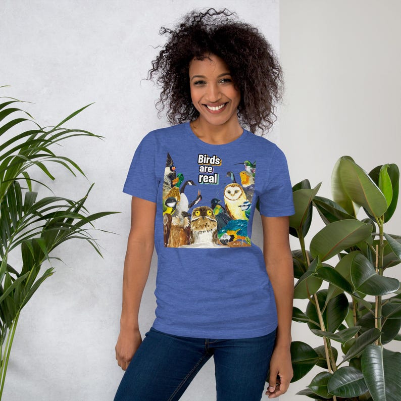 May include: Blue short-sleeve t-shirt with a colorful illustration of various birds. The text "Birds are real" is printed in white. The shirt is made of a soft material.