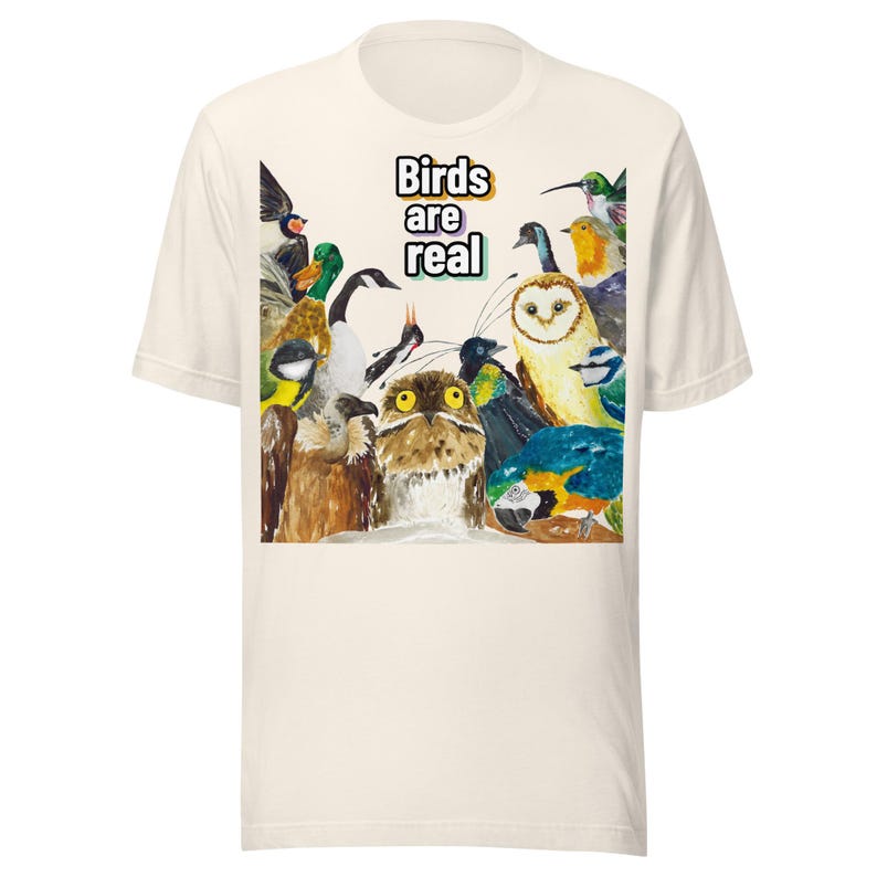 May include: Cream-colored t-shirt featuring a colorful watercolor illustration of various birds. The phrase "Birds are real" is printed in a black and white font above the birds. The shirt is made of a soft, comfortable material.