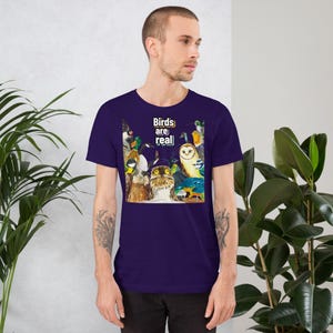 May include: A short-sleeved, dark purple t-shirt featuring a colorful illustration of various birds. The graphic includes owls, ducks, and other avian species. The text "Birds are real" is printed above the birds in white and yellow.