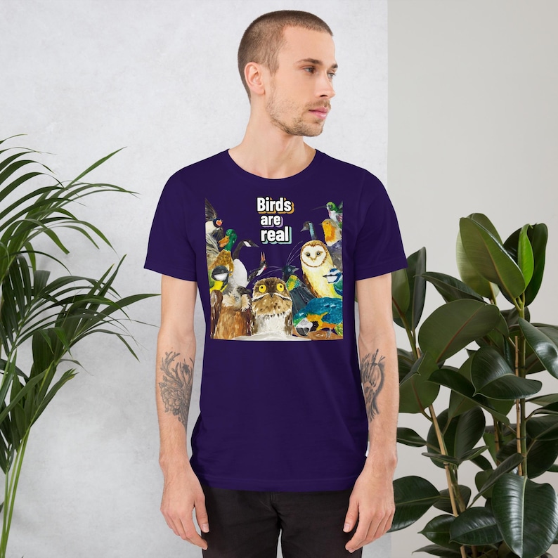 May include: A short-sleeved, dark purple t-shirt featuring a colorful illustration of various birds. The graphic includes owls, ducks, and other avian species. The text "Birds are real" is printed above the birds in white and yellow.