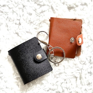 May include: Two small wallets in black and tan, each with a snap closure. One wallet is attached to a silver keyring. The wallets are displayed on a white, textured surface.