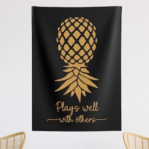 May include: A black and gold tapestry with a pineapple design and the text "Plays well with others".