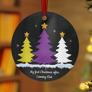 May include: Round Christmas ornament with a black background, featuring three stylized Christmas trees in yellow, purple, and white. The text "My first Christmas after Coming Out" is printed on the bottom. A red ribbon is attached for hanging.