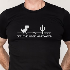 Może przedstawiać: Czarny T-shirt z białym pikselowanym dinozaurem i kaktusem. Pod grafiką znajduje się napis "OFFLINE MODE ACTIVATED".