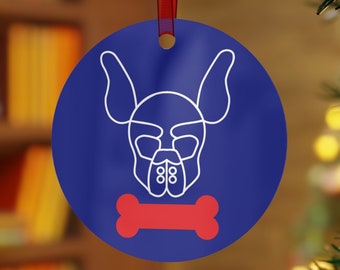 May include: A round, blue Christmas ornament with a white line drawing of a dog's face and a red bone. The ornament hangs from a red ribbon against a blurred background of a Christmas tree and a bookshelf.