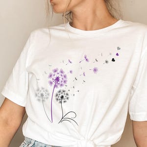 Pode incluir: T-shirt branco com um design de dente-de-leão em roxo e preto. O desenho mostra um dente-de-leão com sementes a voar e formas de coração. Uma peça casual para o dia a dia.