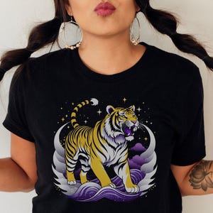 Może przedstawiać: Czarny t-shirt z wystylizowaną ilustracją tygrysa. Tygrys ma żółto-czarne paski, fioletowe akcenty i gwiezdne tło. Projekt jest nadrukowany na czarnym t-shircie z okrągłym dekoltem.