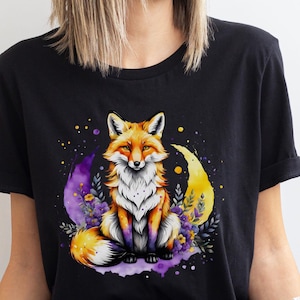 Könnte beinhalten: Schwarzes T-Shirt mit einer Aquarellillustration eines Fuchses, der auf einem lila Halbmond mit gelben Akzenten sitzt. Der Fuchs hat orange-weißes Fell, umgeben von lila Blumen und gelben Punkten.