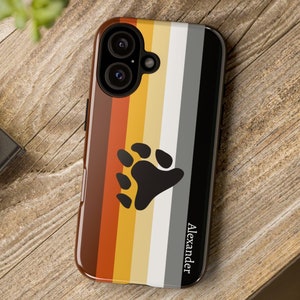 Könnte beinhalten: Eine Handyhülle mit einem Bärenpfotenabdruck auf einem Regenbogen-Pride-Flaggenhintergrund. Die Hülle ist schwarz und hat den Namen "Alexander" unten aufgedruckt.