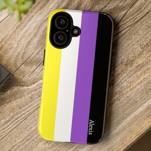 May include: A smartphone case featuring the non-binary pride flag design. The case has a yellow, white, purple, and black striped pattern. The name "Alexis" is printed vertically in white on the black stripe. The case is on a wooden surface.