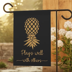 May include: Black garden flag with a gold pineapple design and the phrase "Plays well with others." The flag is displayed on a black metal stand in a garden setting, surrounded by greenery and white flowers.