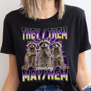 Peut inclure: T-shirt noir avec un graphique de trois ratons laveurs et le texte "They/Them causing May/Hem". Les ratons laveurs sont debout en rang et ont des éclairs violets et jaunes derrière eux.
