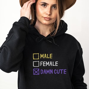 Op de afbeelding: Zwarte hoodie met een gele en paarse afbeelding met de tekst "Male", "Female" en "Damn Cute", waarbij de laatste optie is aangevinkt.