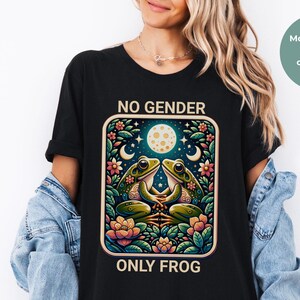 May include: A black t-shirt featuring a graphic print of two frogs sitting among colorful flowers and foliage under a moon and stars. The text NO GENDER is printed above the graphic, and the text ONLY FROG is printed below the graphic.
