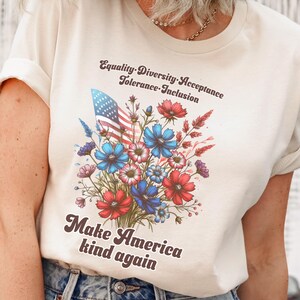 Könnte beinhalten: Ein weißes T-Shirt mit einem Motiv eines Straußes roter, weißer und blauer Blumen mit einer amerikanischen Flagge. Der Text "Equality. Diversity. Acceptance. Tolerance. Inclusion." befindet sich über den Blumen und der Text "Make America kind again" unter den Blumen.