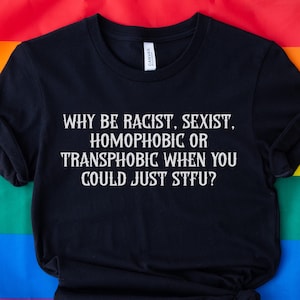 Może przedstawiać: Czarna koszulka z białym napisem "WHY BE RACIST, SEXIST, HOMOPHOBIC OR TRANSPHOBIC WHEN YOU COULD JUST STFU?". Koszulka jest prezentowana na tęczowej fladze.
