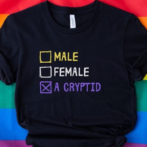 Pode incluir: Camiseta preta com um gráfico branco que diz "Male", "Female", e "A Cryptid" com uma marca de verificação ao lado da última opção.