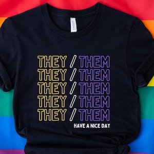 May include: Black t-shirt with the text "THEY/THEM" repeated five times in yellow and purple outlines. The text "HAVE A NICE DAY" is printed below the repeated text.