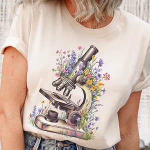 May include: Cream-colored t-shirt featuring a watercolor illustration of a vintage microscope surrounded by colorful wildflowers. The microscope is detailed with silver and black accents, and the flowers include purple, yellow, and pink blooms.