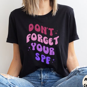 May include: A black t-shirt with the text "Don't forget your SPF" in a gradient of pink, purple, and blue. The text is in a retro font style.