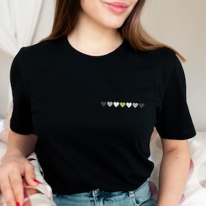 May include: Black t-shirt with a row of small hearts in shades of gray, white, and green. The green heart is in the center of the row.