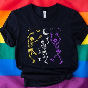 May include: Black t-shirt featuring a graphic of three dancing skeletons in yellow, white, and purple. The design includes a crescent moon, stars, and bats. The shirt is displayed on a rainbow flag.