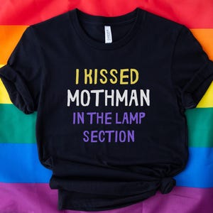 May include: Black t-shirt with the text "I kissed Mothman in the lamp section" in yellow and purple lettering. The shirt is displayed on a rainbow pride flag.