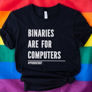 May include: A black t-shirt with the text "Binaries are for computers" in white lettering and the hashtag "#PROUDENBY" below. The t-shirt is folded and lying on a rainbow pride flag.
