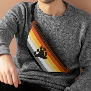May include: A black fanny pack with a brown, yellow, white and grey striped pattern and a black bear paw print. The fanny pack is being worn by a person.