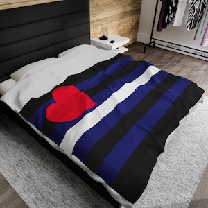 Op de afbeelding: Een wit bed met een zwart, blauw en wit gestreept deken met een rood hart in het midden. Het deken is gemaakt van zachte, comfortabele stof en perfect om te knuffelen op de bank of in bed.