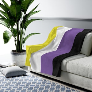 May include: A soft, plush throw blanket featuring the non-binary pride flag colors: yellow, white, purple, and black. The blanket is draped over a white couch with a green plant in a pot to the left.