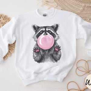 May include: White sweatshirt with a black and white illustration of a raccoon blowing a pink bubble.