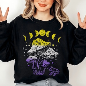May include: Black sweatshirt with a graphic of purple and yellow mushrooms with a crescent moon design and a frog. The text "Shroom Queens" is printed on the shirt.