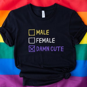 May include: Black t-shirt with a white box checked with a purple X and the text "DAMN CUTE" below it. Above the checked box are two unchecked boxes with the text "MALE" and "FEMALE" in white.