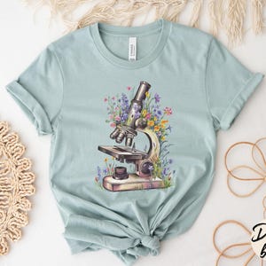 May include: Dusty blue t-shirt featuring a detailed illustration of a vintage microscope surrounded by colorful wildflowers. The shirt is made of soft material and has a classic crew neck design. The word "Dusty blue" is written in a script font.