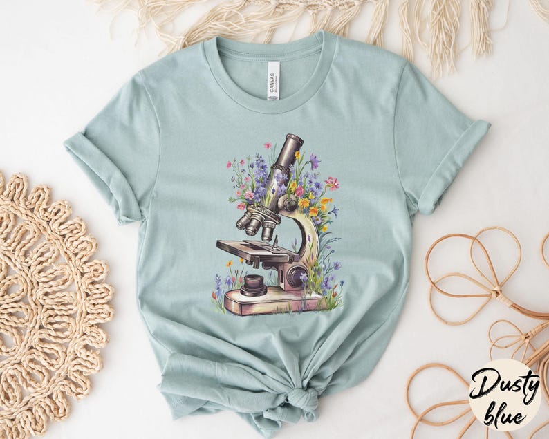 May include: Dusty blue t-shirt featuring a detailed illustration of a vintage microscope surrounded by colorful wildflowers. The shirt is made of soft material and has a classic crew neck design. The word "Dusty blue" is written in a script font.