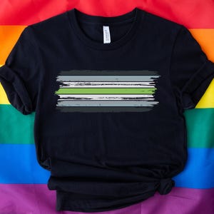 May include: Black t-shirt with a horizontal brushstroke design in grey, green, and white. The shirt is made of soft material and is displayed on a rainbow flag background. The design is centered on the chest.