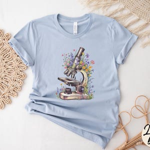 May include: Light blue t-shirt featuring a watercolor illustration of a vintage microscope surrounded by colorful wildflowers. The shirt has a classic crew neck and short sleeves. The words "Light Blue" are written in a cursive font on the bottom right corner.