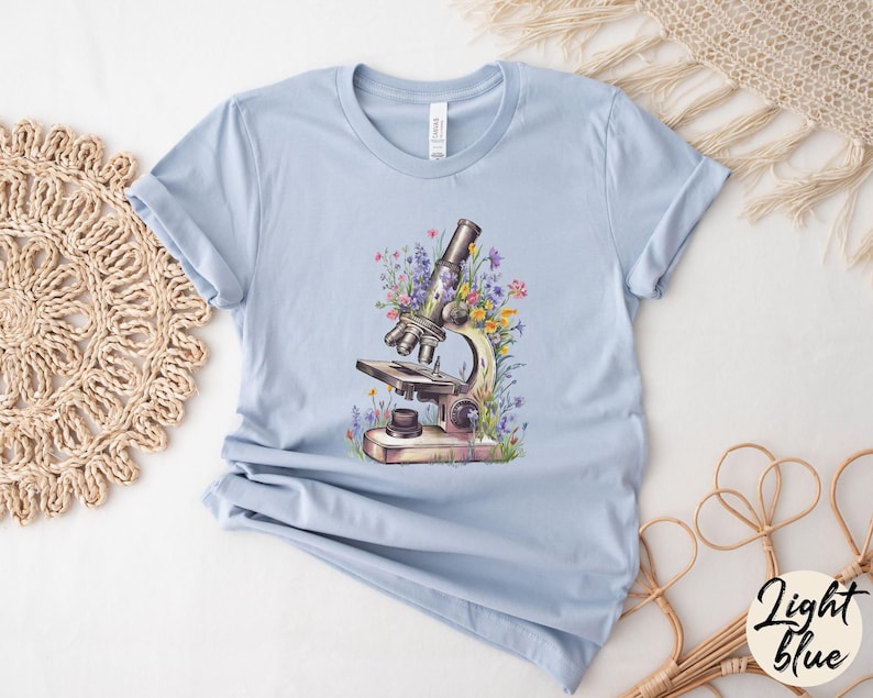 May include: Light blue t-shirt featuring a watercolor illustration of a vintage microscope surrounded by colorful wildflowers. The shirt has a classic crew neck and short sleeves. The words "Light Blue" are written in a cursive font on the bottom right corner.