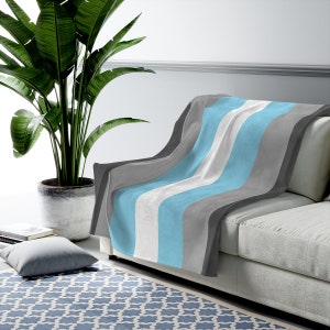 May include: A soft, striped throw blanket in shades of gray, white, and light blue. The blanket is draped over a gray couch.