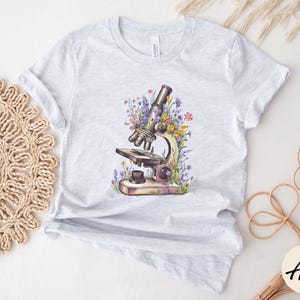 May include: Light gray t-shirt featuring a watercolor illustration of a vintage microscope surrounded by colorful wildflowers. The microscope is detailed with brown and silver tones, and the flowers include purple, yellow, and pink hues.