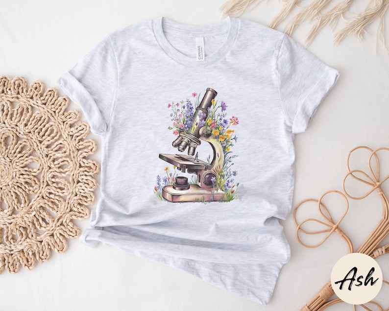 May include: Light gray t-shirt featuring a watercolor illustration of a vintage microscope surrounded by colorful wildflowers. The microscope is detailed with brown and silver tones, and the flowers include purple, yellow, and pink hues.