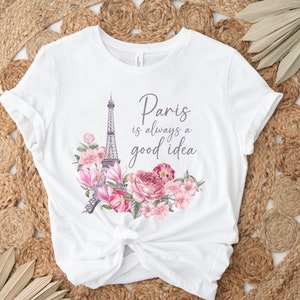 May include: White t-shirt with a design featuring the Eiffel Tower and pink and purple flowers. The text "Paris is always a good idea" is written in a script font. The shirt is knotted at the waist.
